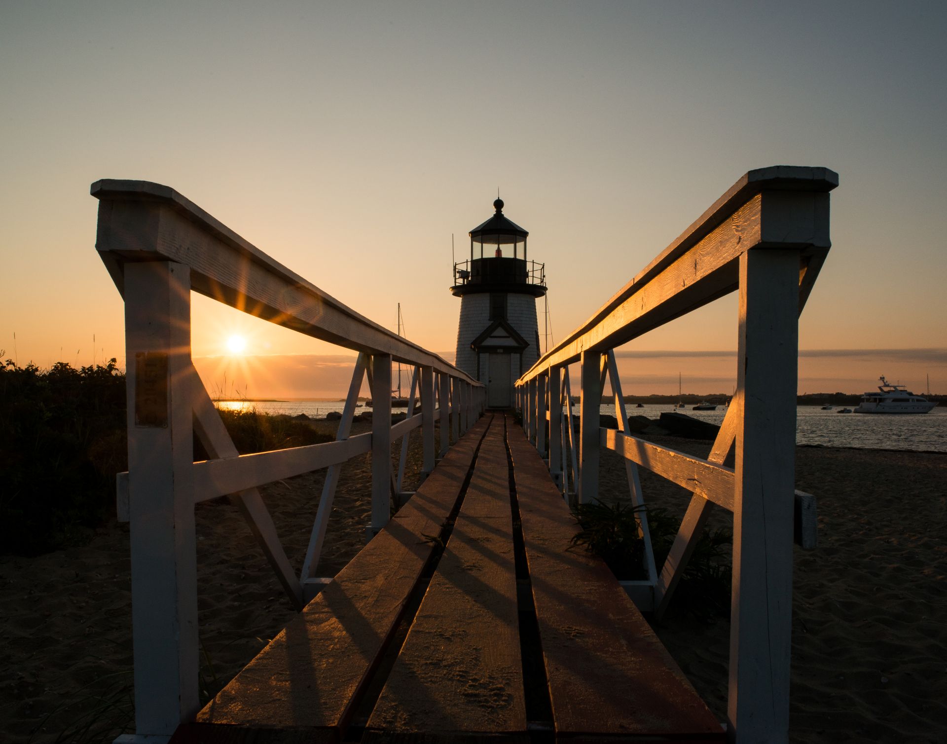 Sunrise illuminates a wooden walkway leading to a white lighthouse, with tranquil waters and boats in the background, capturing coastal serenity.
