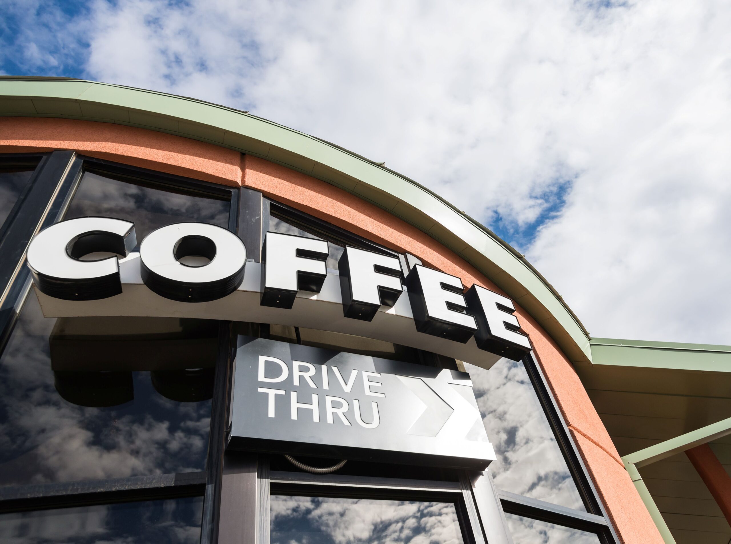 Coffee shop signage highlights the drive-thru option. The architecture features a curved roof and glass windows reflecting a partly cloudy sky.
