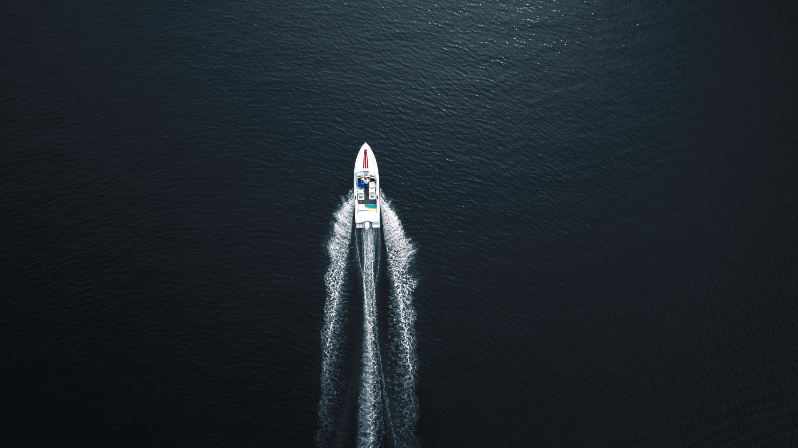 Aerial top-down photo of a white motorboat cruising through deep dark blue water, leaving a dynamic V-shaped wake behind. The minimalistic composition emphasizes the contrast between the boat and the